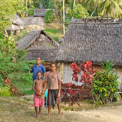 Three children stand in front of traditional thatched-roof houses in a Sepik River village, Papua New Guinea.