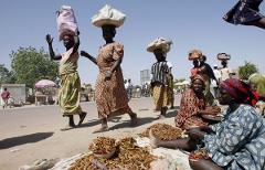 A group of women in patterned dresses walk and sit on the side of a road, some carrying bags on their heads, while others sell dried goods.
