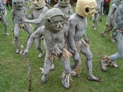 Goroka Day Tour A group of people in traditional Asaro Mudmen costumes, with grey clay covering their bodies and wearing masks, are standing on a grassy field.