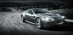 A silver Aston Martin sports car speeds around a curving road, the motion blurring its wheels against a grayscale landscape.