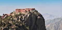 A salmon-colored building perched atop a rocky mountain peak with layered mountains fading in the background under a clear blue sky.