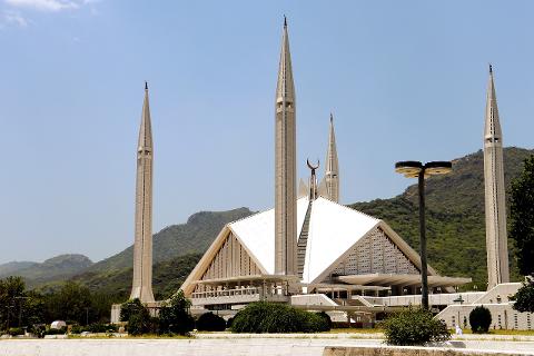 The image shows the Faisal Mosque in Islamabad, Pakistan, a large modern mosque with distinctive minarets and a triangular prayer hall, set against a backdrop of green hills.