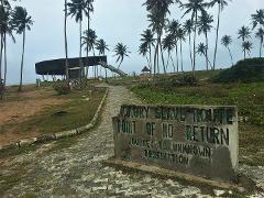 A sign reading "Badagry Slave Route Point of No Return" stands at the beginning of a stone path leading towards a structure and palm trees against a cloudy sky.