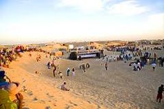 Here's a concise description of the image:A wide shot of a desert music festival scene with crowds gathered around a stage set up on the sand under a clear sky.