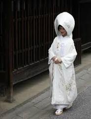 A Japanese bride in a traditional white kimono and wataboshi hood walks on a stone path next to a dark wooden fence.