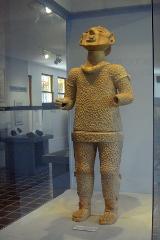 A full-bodied earthen sculpture of a human figure, in a display case, featuring a textured surface resembling scales or chainmail.