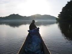 A person is seen paddling a small boat on a calm river, with misty hills in the background under a bright sky.