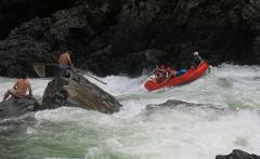 Colombia River Tour: Samana and Verde A group of people in an orange raft are navigating through rapids as two shirtless men sit on rocks nearby.