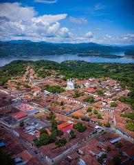 An aerial view showcases a town with red-tiled roofs nestled among lush greenery, a lake, and mountains under a bright blue sky with scattered clouds.