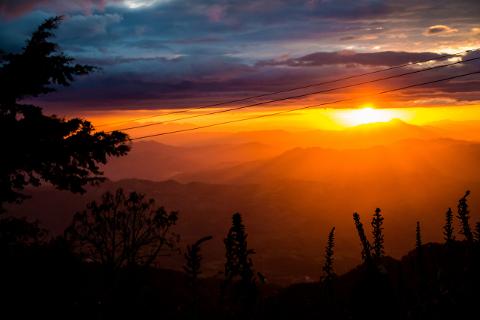 A vibrant sunset bathes a mountain range in golden light, with silhouettes of trees and plants in the foreground and power lines stretching across the sky.