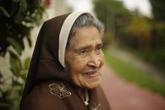An elderly woman, likely a nun, is captured in a warm, close-up shot, wearing a brown habit with a white trim and embroidered butterfly detail, with a gentle smile on her face.