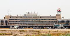 A full shot of Baghdad International Airport on a clear day, featuring the main building, the control tower with a red and white spherical top, and several airplanes on the tarmac.