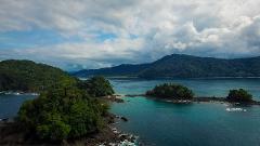 An aerial view showcases lush, green islands in turquoise water, under a cloudy sky, revealing a serene tropical coastal landscape.