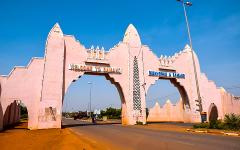 A pink, ornate gate welcomes visitors to Bamako, with "Welcome to Bamako" on one side and "Bienvenue a Bamako" on the other, under a clear blue sky.