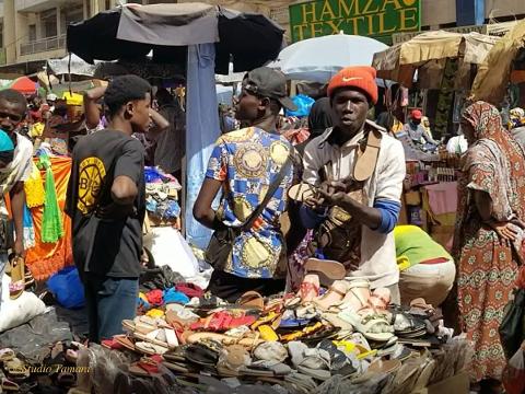 Full Day Tour To Siby And Bamako A vendor in a red beanie holds up sandals at a bustling market stall filled with footwear in Bamako, Mali.