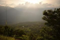 A scenic view of a lush, green valley under a cloudy sky with sun rays breaking through, framed by vegetation in the foreground.