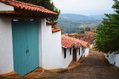 A picturesque street scene with a turquoise door, white walls, and terracotta roofs leading towards a view of mountains and a church in Barichara, Colombia.