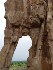A large rock formation with an arch-shaped opening frames a distant green landscape under a cloudy sky.