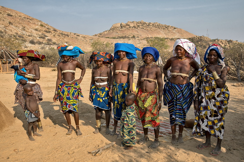 A group of Himba women and children stand in a desert landscape, adorned with colorful fabrics and traditional jewelry.