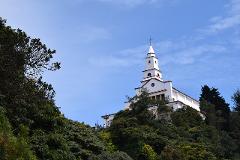 A white church with a red trim sits atop a green, tree-covered hill, framed against a blue sky with wispy clouds.