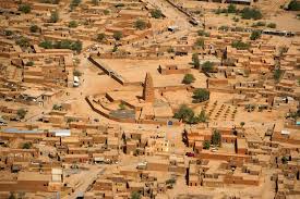 Certainly! Here's a description of the image within the character limit:An aerial view shows a dense, mud-brick city with a prominent mosque at its center, characteristic of Sahelian architecture in a dry, dusty landscape.