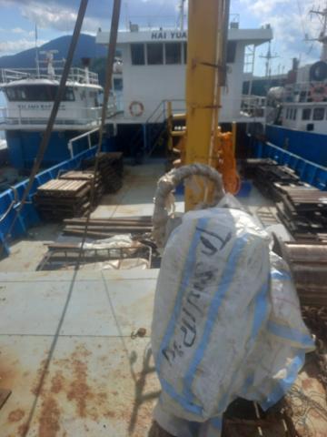 The deck of a ship or boat is filled with supplies and equipment, including pallets, pipes, and a large bag with writing on it, while other boats are docked alongside.