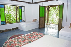 Birding - Cali, Coffee Region & Medellin The image shows a bright, airy bedroom with white walls and blue floors, featuring two beds, a window with colorful glass panes, and an open doorway leading to a lush, green outdoor area.