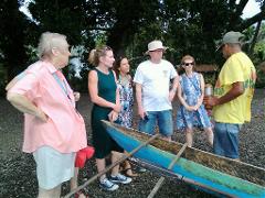 A tour group stands around a traditional canoe, listening to a guide, likely on a beach in Alotau.