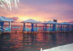 Here's a description of the image within the character limit:Several blue-trimmed cabanas line a pier over water reflecting a vibrant sunset sky in Bocas del Toro.