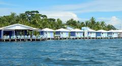 A row of charming blue and white bungalows perched on stilts above a shimmering turquoise sea, framed by lush tropical vegetation and a vibrant blue sky.