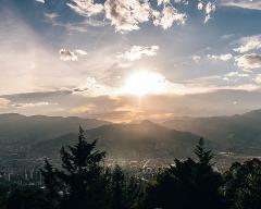 A scenic view of a city nestled in a valley surrounded by mountains, bathed in the golden light of a setting sun, partially obscured by clouds and framed by silhouetted trees.