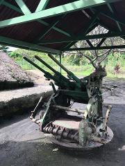An old, green, triple-barreled anti-aircraft gun is displayed under a covered structure in an outdoor setting.