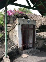 An old concrete bunker with a rusty metal gate and a blue anchor symbol is set into the side of a rocky hill with vibrant pink flowers growing on top.