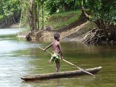 A young boy, seemingly from a local tribe, stands in a small dugout canoe on the Sepik River, holding a paddle, likely navigating its waters.