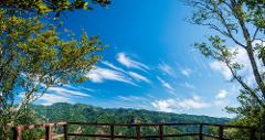 A scenic view from a wooden platform showcases lush green mountains under a vibrant blue sky with wispy clouds, framed by trees on either side.