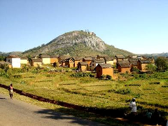 A village in Madagascar sits at the base of a large, rocky mountain, with rice paddies in the foreground.