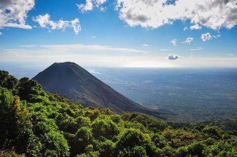 A majestic volcano rises from lush green foliage under a bright blue sky with puffy white clouds.