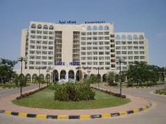 The Libya Hotel Kempinski stands prominently against a clear blue sky, surrounded by green lawns and palm trees.
