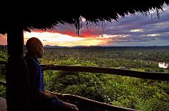From a hut, a person contemplates a vibrant sunset over a vast, lush landscape with mountains in the distance, a river winding through it.