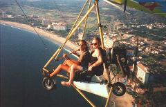 Two men in sunglasses are flying over a beach city in a small ultralight aircraft with an engine.