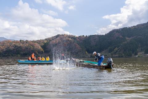 Two boats are on a lake, one with several people in orange and yellow life vests, the other with a man in blue overalls hauling in a net, creating a large splash.
