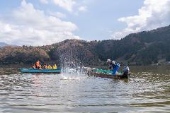 Discover 400 Years of Fishing Tradition on Lake Mikata Two boats are on a lake, one with several people in orange and yellow life vests, the other with a man in blue overalls hauling in a net, creating a large splash.