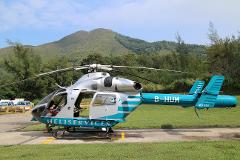 A silver and blue helicopter labeled "HELISERVICES" with the tail number "B-HUM" sits on a pad against a backdrop of green trees and a distant mountain.