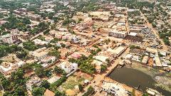 An aerial view shows a dense urban landscape of buildings, trees, and a pond, with a mix of residential and commercial areas under a bright sky.