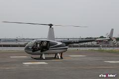 Un helicóptero gris con número de cola JA7936 se encuentra en la pista con las palas del rotor girando, el piloto visible en la cabina abierta y un paisaje urbano borroso en el fondo.
