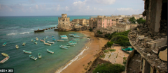 An aerial shot of a harbor and beach dotted with small boats, a rocky shoreline with a large, weathered castle-like structure, and a city skyline marred by bombed-out buildings.