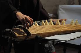 A person plays a Koto, a traditional Japanese stringed instrument, with sheet music nearby.