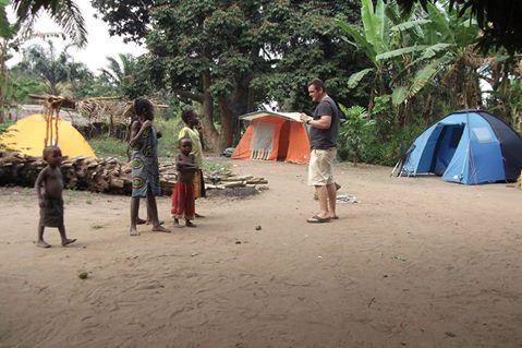 An Caucasian man is seen holding a notebook while talking to three African children in front of multiple tents on a dirt ground.
