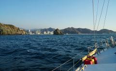 Sailing the Caribbean in Colombia A sailboat sails on the ocean towards the land, which is composed of cliffs, buildings, and mountains.
