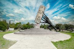A bronze statue of a group of men holding up a cylinder with symbols on it stands on a tiered platform in a park with a blue sky and white clouds.